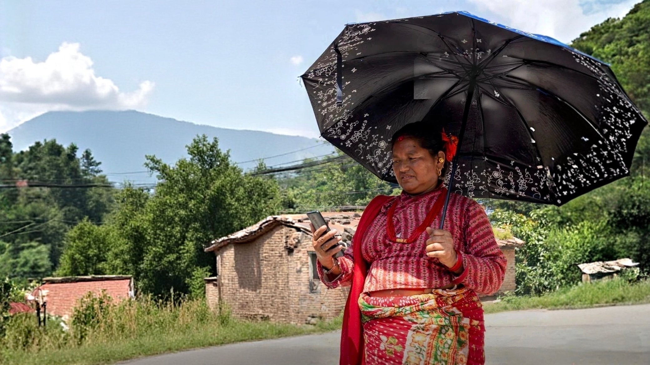 A woman in traditional attire holds a large black umbrella and looks at her phone, standing by a rural roadside with green trees, old brick houses, and a mountain in the background under a partly cloudy sky.