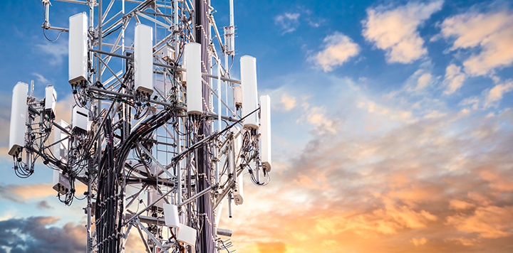 A close-up view of a telecommunications tower with multiple white antennas and cables, set against a vibrant sky with scattered clouds and a warm, colorful sunset in the background.