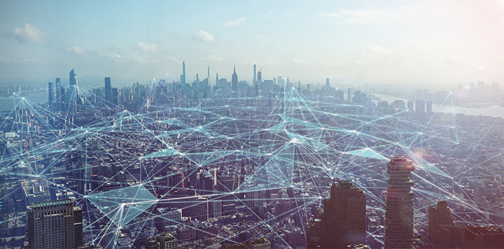 A city skyline with tall buildings under a hazy sky. Digital blue lines and nodes are superimposed over the city, symbolizing connectivity, technology, and communication networks throughout the urban landscape.