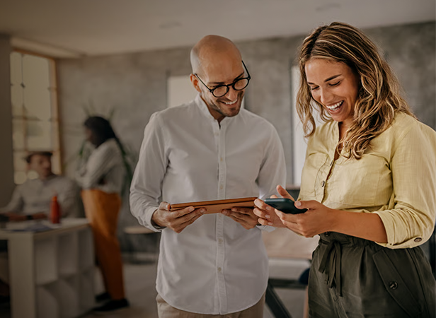 Two colleagues stand together in an office, smiling and looking at digital devices. The man holds a tablet, whilst the woman looks at her mobile. Two other colleagues converse blurred in the background. The atmosphere appears casual and collaborative.