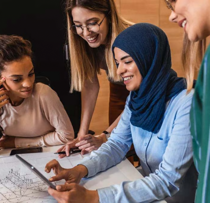 Four women gather around a table, smiling and collaborating. One woman in a blue hijab holds a tablet, while another points at a printed diagram. They appear engaged and focused, suggesting teamwork and shared ideas in a modern office setting.