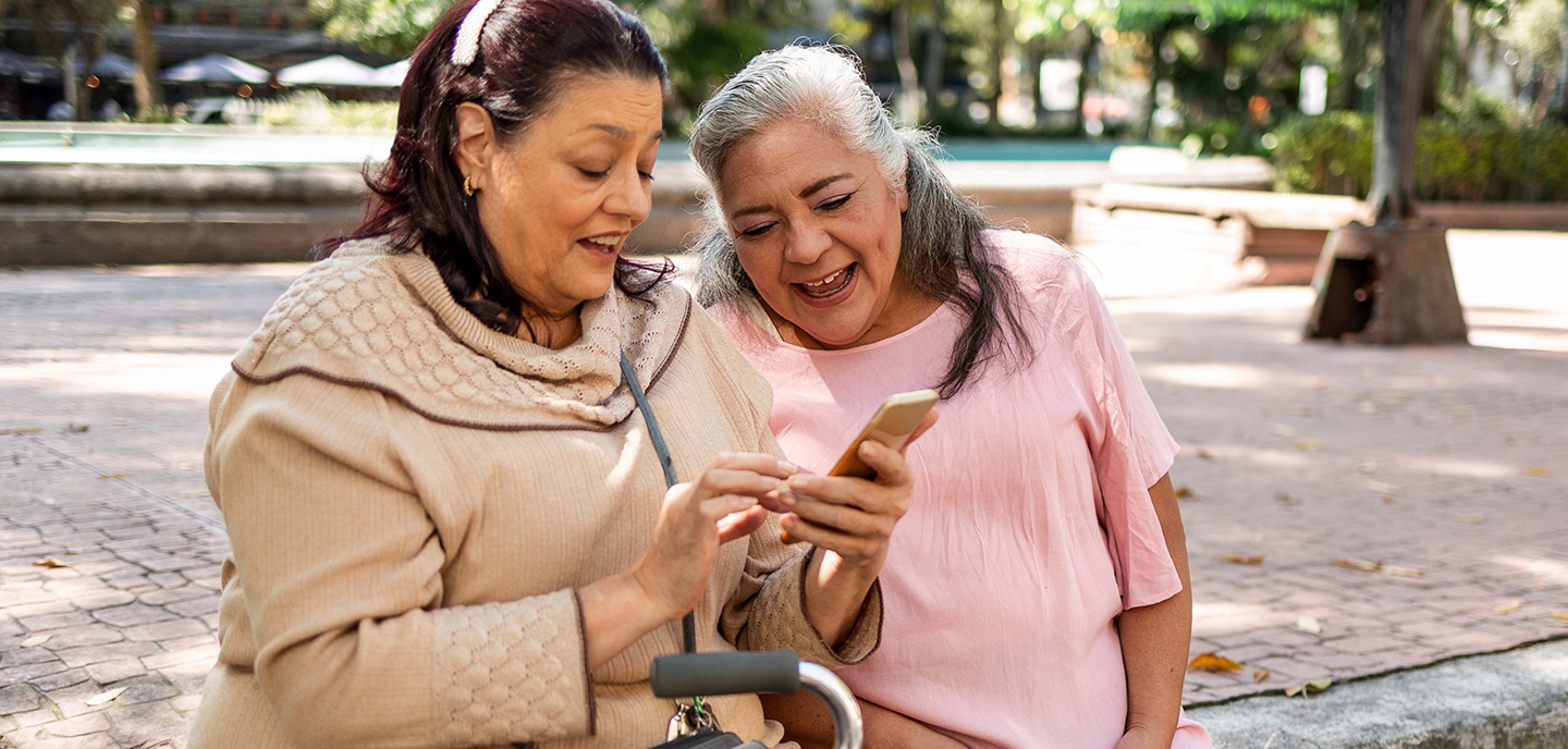 Two older women sit close together on a park bench, smiling and looking at a mobile phone one is holding. One woman wears a light beige jumper and has a walking stick, while the other wears a pink dress. Trees and sunlight fill the background.