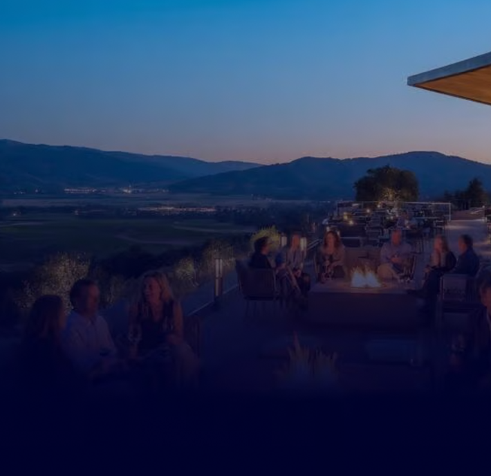 People sit around outdoor fire pits on a terrace at dusk, overlooking a scenic valley and distant mountains under a clear evening sky. The atmosphere is relaxed, with groups enjoying conversation and drinks as lights begin to glow in the background.