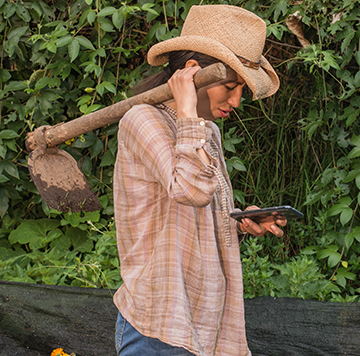 A person in a straw hat and checked shirt holds a muddy hoe over their shoulder while looking at a mobile phone. They are standing outdoors in front of dense green foliage, combining gardening with using technology.