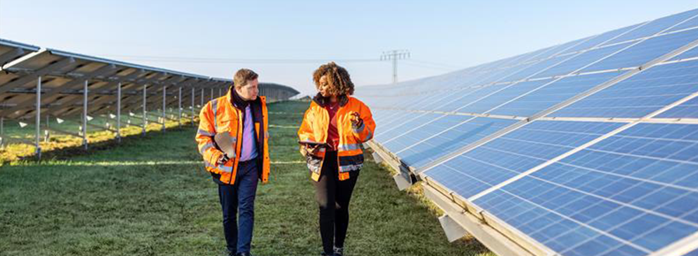 Two people in orange safety jackets and holding clipboards walk beside rows of solar panels outdoors. They appear to be inspecting or discussing the solar installation on a grassy field under a clear blue sky.