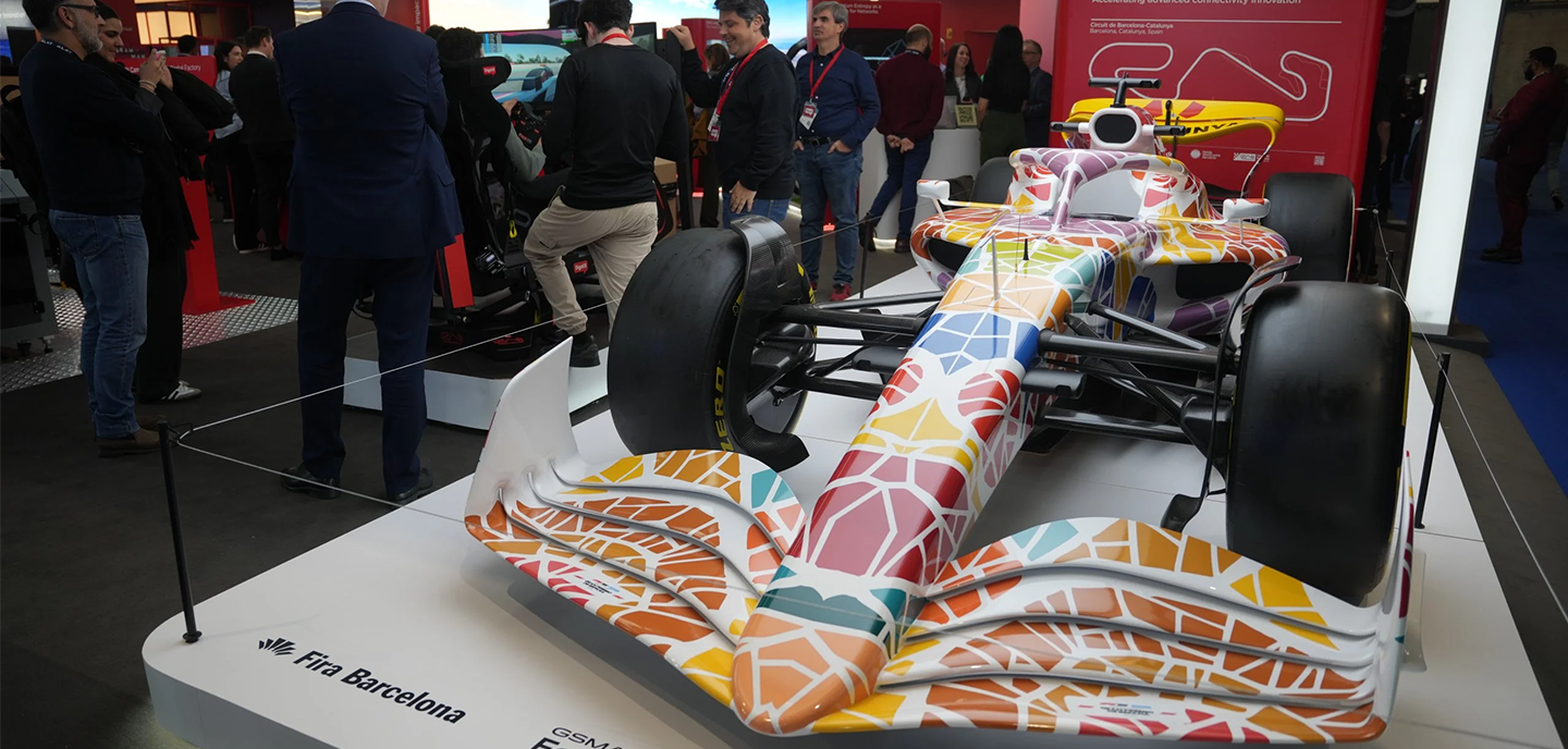 A Formula 1 car with a colourful mosaic pattern is displayed on a white platform labelled "Fira Barcelona." People stand and observe the car in a busy exhibition hall, with red signs and informational stands in the background.