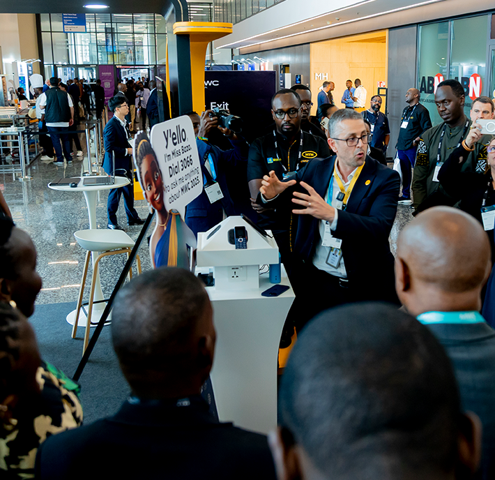 A group of people gathers around a man in a grey suit and yellow button, who is animatedly presenting at an indoor exhibition. Attendees listen closely. Colourful banners, stands, and a “Y’ello” sign are visible in the background, indicating a tech or business event.