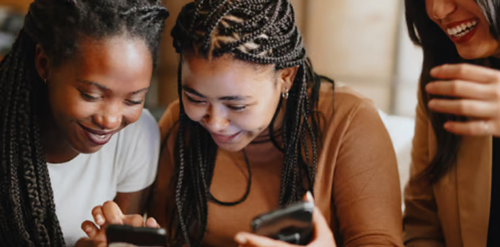 Three women sit closely together, smiling and laughing whilst looking at mobile phones. One woman wears a white shirt, another a brown shirt with plaits, and the third is partly visible, wearing a beige jacket. They appear to be enjoying each other's company.