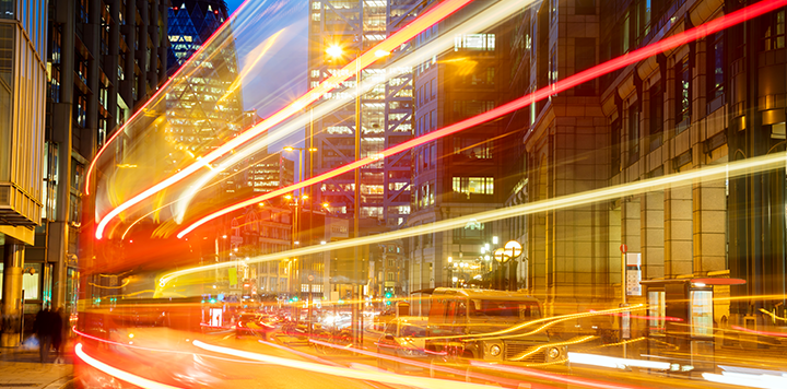 A city street at night with tall, illuminated office buildings. Colourful light trails from moving vehicles streak across the scene, creating dynamic lines of red, yellow, and white, highlighting the energy and movement of urban nightlife.