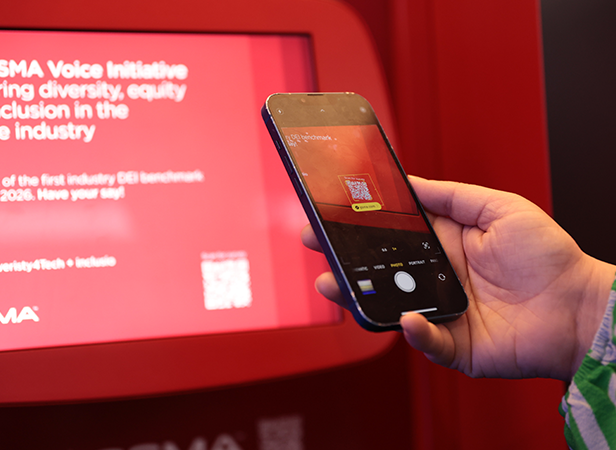 A person holds a mobile phone, scanning a QR code on a red digital display. The phone’s screen clearly shows the QR code in focus. The display behind promotes diversity, equity, and inclusion in the tech industry, with white text on a red background.