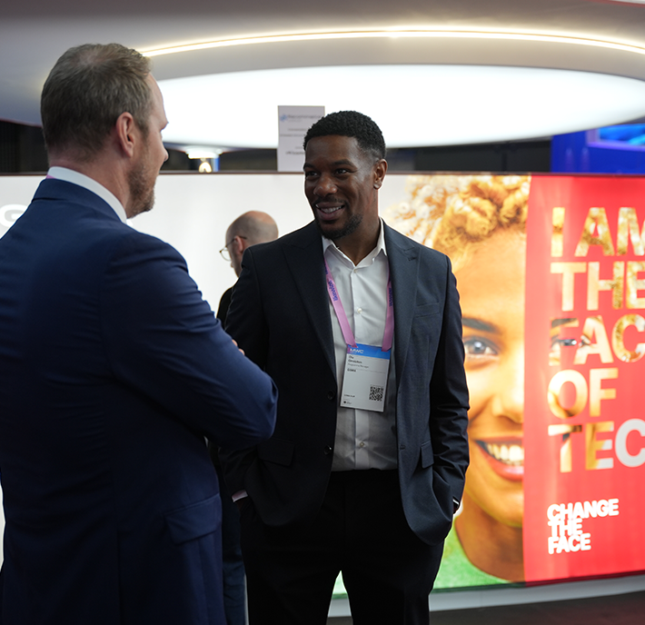 Two men in suits are having a conversation at an event. One faces the camera and smiles, wearing a badge. Behind them is a bright display with a woman's face and text reading “I AM THE FACE OF TECH” and “CHANGE THE FACE.”.