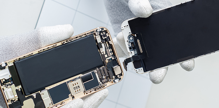 A person wearing white gloves is holding a mobile phone that is opened, separating the screen from the body. The internal components, battery, and circuit board of the phone are visible. The background is out of focus and has a white tiled surface.