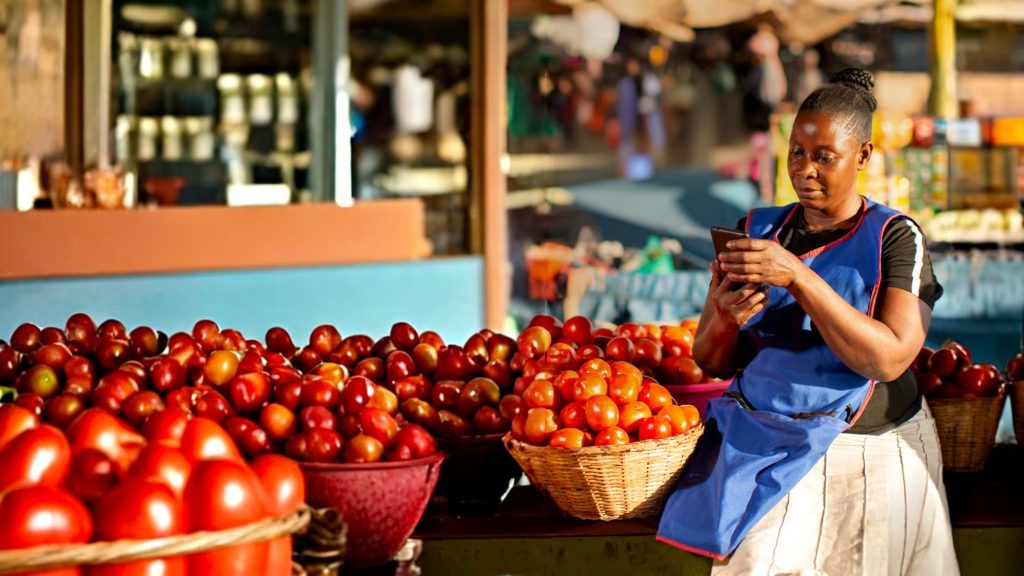 A woman vendor in a blue apron stands at a market stall filled with baskets of tomatoes, looking at her mobile phone. Sunlight highlights the vibrant red tomatoes, and shelves with jars are visible in the blurred background.