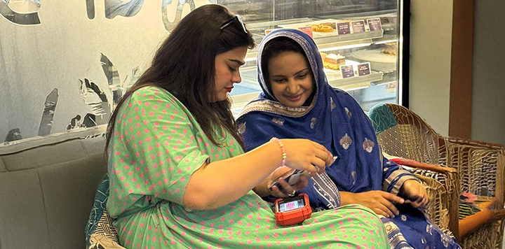 Two women sitting on wicker chairs indoors, smiling and looking at a mobile phone. One woman wears a green dress with pink spots, the other wears a blue patterned headscarf and dress. Behind them is a display case with bakery items.