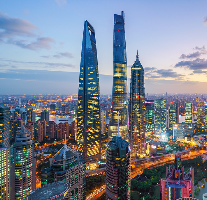 Aerial view of Shanghai’s modern cityscape at dusk, featuring the iconic Shanghai Tower, Shanghai World Financial Centre, and Jin Mao Tower surrounded by illuminated skyscrapers and busy streets under a blue and purple sky.