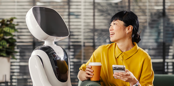 A smiling woman in a yellow shirt sits holding a coffee cup and mobile, looking at a sleek, white humanoid robot with a blank screen face. They appear to be interacting in a modern, sunlit office setting with large windows.