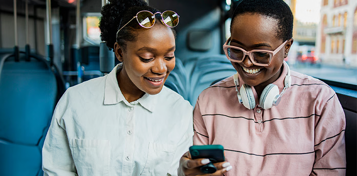 Two smiling people sit on a bus, looking at a mobile. One wears a white shirt and sunglasses on her head. The other wears a pink striped shirt, large glasses, and headphones round her neck. Blue bus seats are visible in the background.