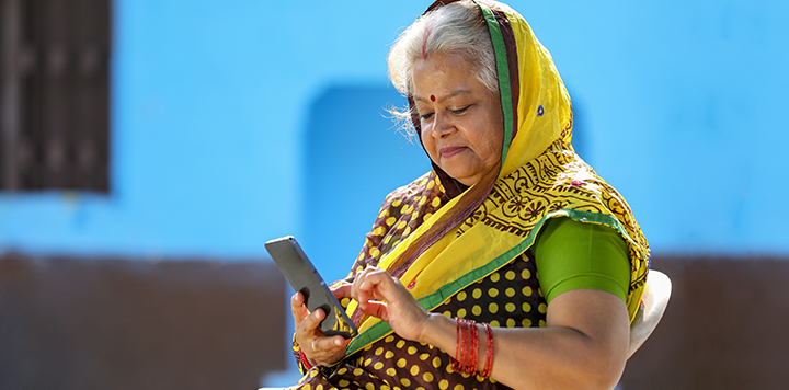 An older woman wearing a colourful saree with a yellow and green scarf sits outdoors, focused on using a mobile phone. She has grey hair, wears bangles, and is sitting in front of a bright blue wall with sunlight illuminating her face.