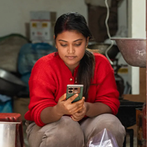 A woman wearing a red jumper sits indoors, looking at a mobile phone in her hands. She appears focused. There are various bags, containers, and kitchen items around her, suggesting a busy or work-related environment.