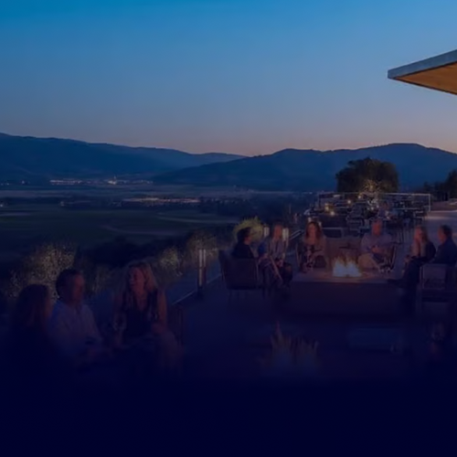 People sit around outdoor fire pits on a patio in the evening, overlooking a scenic valley with mountains in the distance. The sky is clear and blue, and the landscape is softly lit, creating a relaxing and inviting atmosphere.
