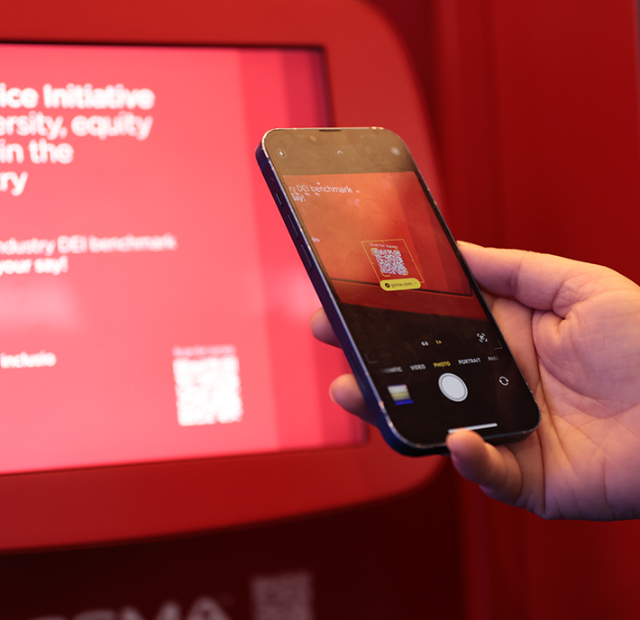 A hand holds a mobile phone, scanning a QR code on a red display screen. The display shows blurred white text about diversity, equity, and inclusion. The phone camera screen clearly focuses on the QR code, with the yellow scanner frame visible.