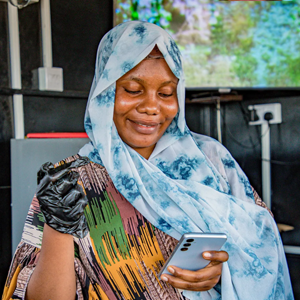 A woman wearing a patterned blue and white headscarf and colourful dress smiles whilst looking at her mobile phone. She holds a tool in her gloved hand. The background shows electrical sockets and a blurred screen displaying greenery.