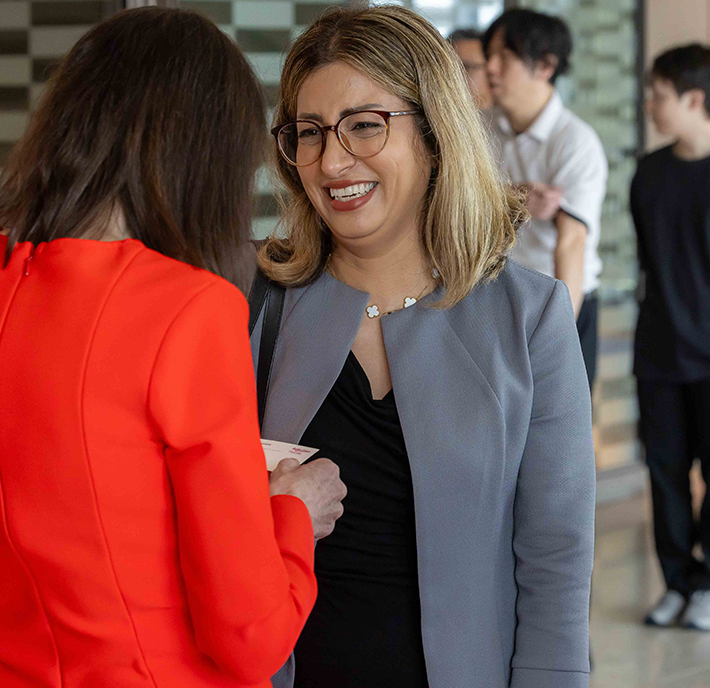 A woman with glasses and shoulder-length blonde hair, wearing a grey blazer over a black top, smiles whilst talking to another woman in a bright red outfit. Two people stand in the blurred background in a modern indoor setting.