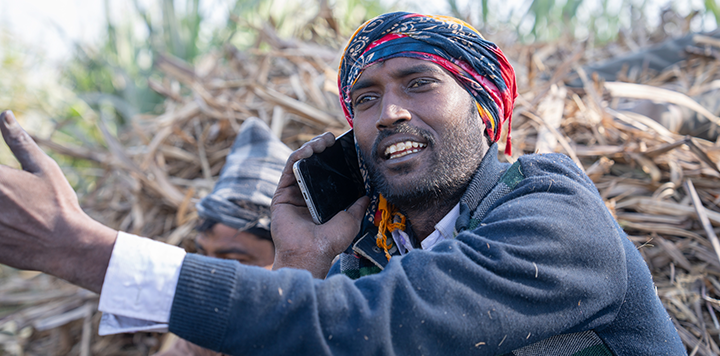 A man wearing a colourful headscarf and dark jumper is talking on a mobile phone. He is gesturing with his left hand and appears to be sitting outdoors on a pile of dry plants or sugarcane stalks, with greenery in the background.