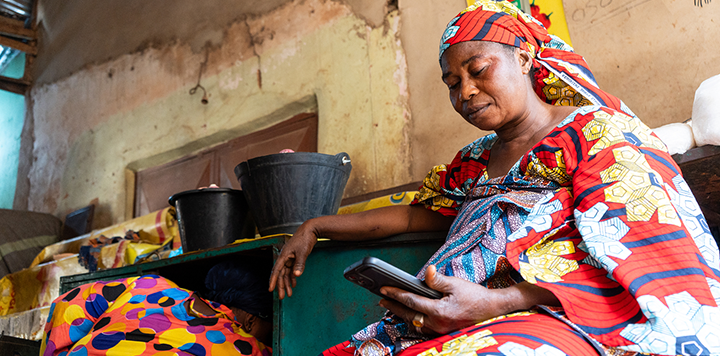A woman in vibrant, patterned traditional clothing and headscarf sits indoors, looking at a mobile phone. Behind her, another person in colourful attire crouches near a green cupboard with black buckets on top. The walls are old and worn.