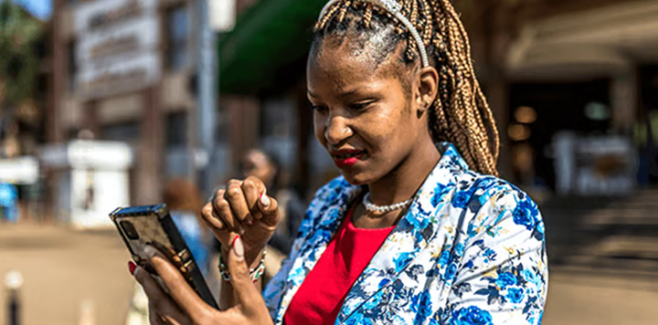 A woman with plaited hair, wearing a blue floral blazer and red top, stands outdoors in bright sunlight. She looks intently at her mobile phone, holding it in one hand while using her other hand to touch the screen. Urban buildings are blurred in the background.