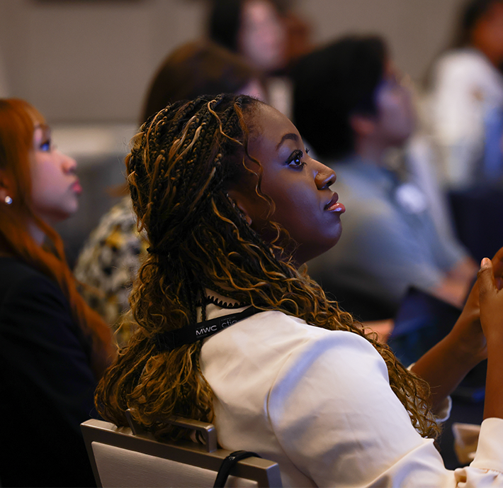 A woman with plaited hair and a white blazer sits attentively among a diverse group of people, all facing forward in a conference or seminar setting, appearing engaged and focused on a presentation out of shot.