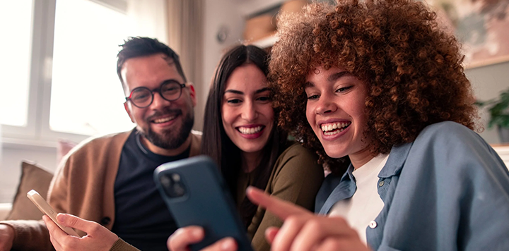 Three young adults sit closely together on a sofa, smiling and laughing as they look at a mobile phone that one person is holding, suggesting they are enjoying something on the screen. Warm indoor lighting creates a cosy atmosphere.