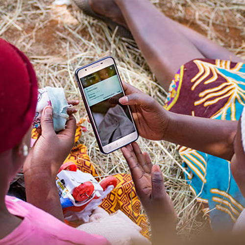 Two people sit on straw-covered ground. One holds a mobile phone displaying a message, while the other holds money and papers. Both wear colourful, patterned clothes. The focus is on their hands and the phone, with their faces out of view.