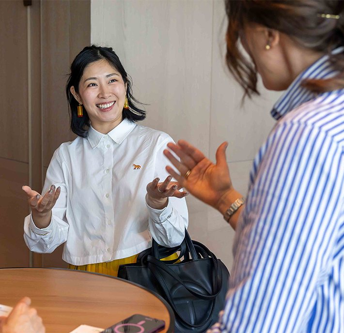 Two women are talking by a round table indoors. One, wearing a white shirt with a small animal badge, is smiling with open hands. The other, in a blue striped shirt, gestures with one hand, her face turned away. A black bag is on the table.