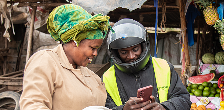 A smiling woman in a green headscarf and beige coat stands beside a man in a motorcycle helmet and reflective vest. They look at a red mobile phone together at an outdoor market with fruit and vegetables visible in the background.