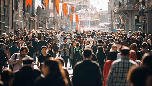A densely packed crowd of people walk along a sunlit city street decorated with red flags. Buildings line both sides, and the street is filled with people of various ages, some in conversations, others moving forward. The atmosphere is lively and bustling.