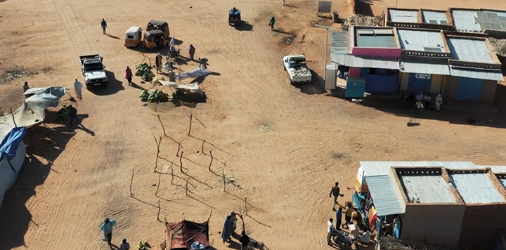 Aerial view of a dusty marketplace with vehicles, scattered stalls, people walking, and small buildings. Some structures have brightly coloured walls and tin roofs. The ground is bare, reddish earth, with small groups gathered near stalls and shelters.