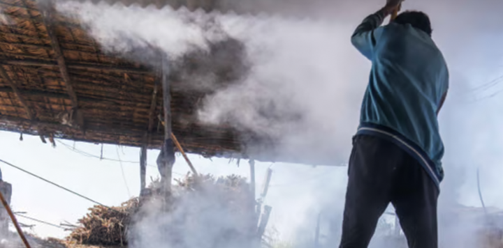 A person stands under a rustic, thatched-roof shelter surrounded by thick white smoke or steam. Their back is turned, and they hold an object raised in one hand. Sunlight filters through the scene, highlighting the texture of the structure.