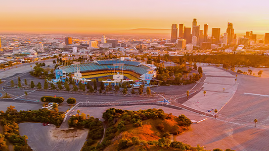 Aerial view of Dodger Stadium in Los Angeles at sunset, with the city skyline in the background. The stadium is surrounded by mostly empty car parks and greenery, while tall buildings rise in the golden-hued distance.