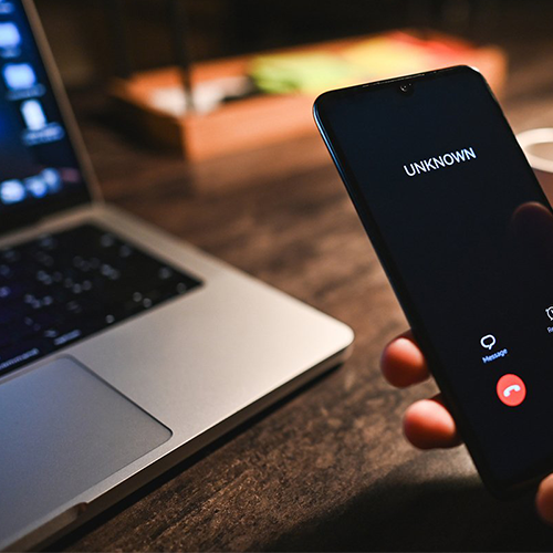 A person holds a mobile phone displaying an incoming call from "UNKNOWN" next to a silver laptop on a wooden desk. The phone screen shows options to message or decline the call. The background is softly lit and slightly blurred.