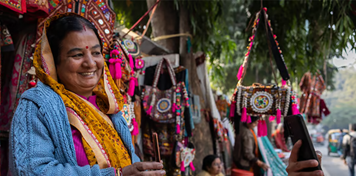 A smiling woman in colourful traditional clothes stands by a market stall selling vibrant embroidered handbags, engaging with someone holding a mobile, with trees and hanging bags in the background.
