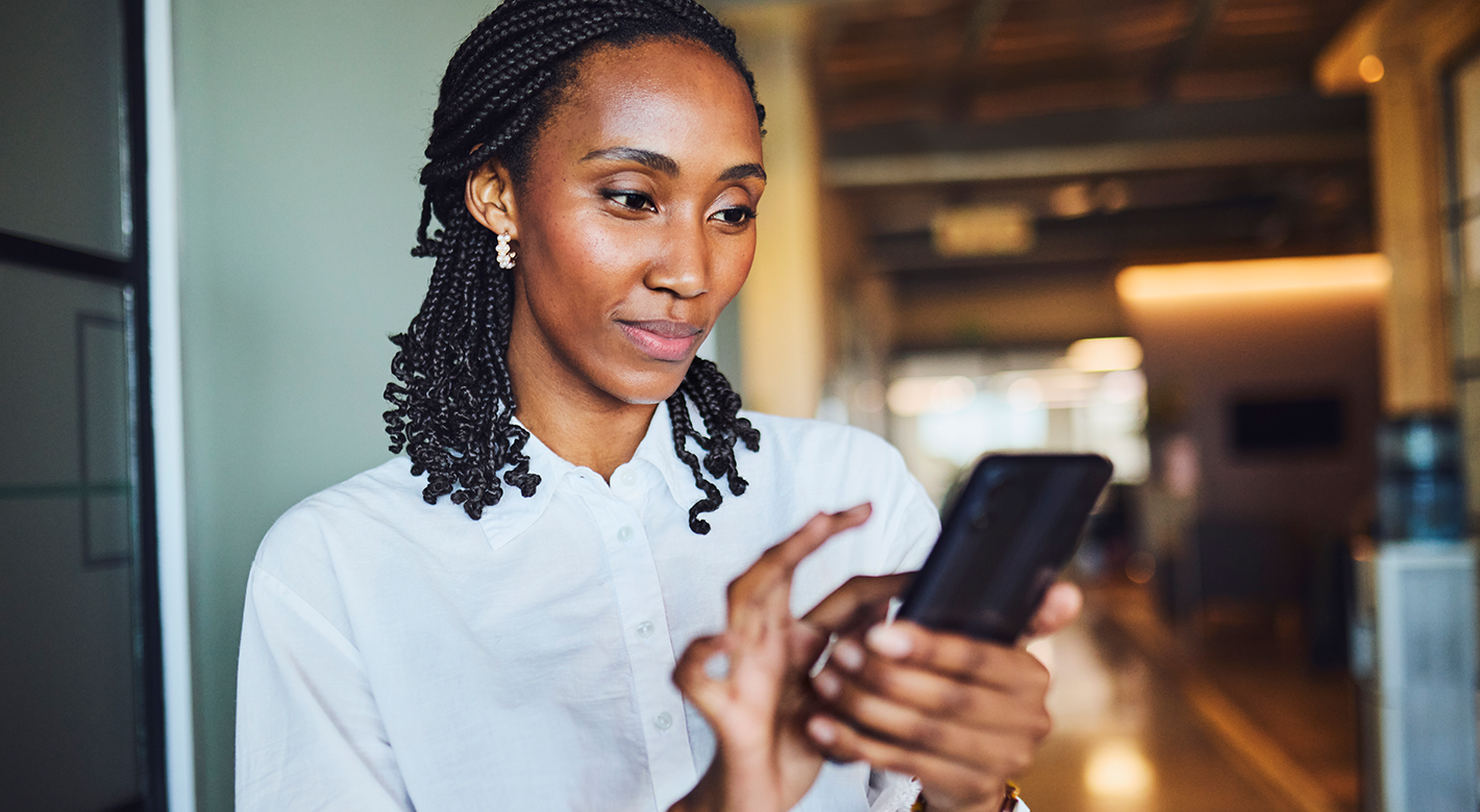 A woman with plaited hair, wearing a white shirt, stands indoors smiling softly whilst using a mobile phone. The background is softly blurred, showing a warm, modern interior space. She appears focused and engaged with her device.