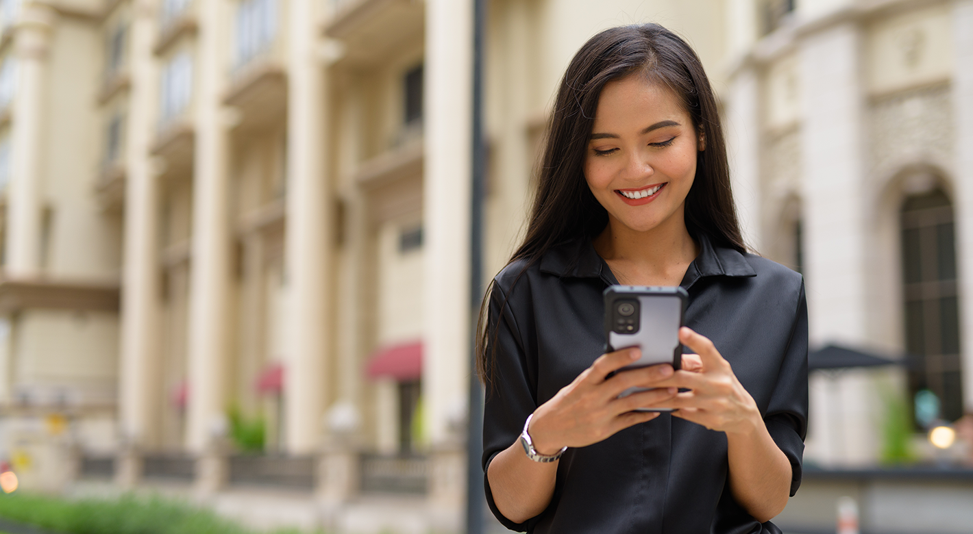 A woman with long dark hair, wearing a black blouse, stands outdoors in front of a beige building, smiling as she looks at her mobile phone. The background is bright and slightly blurred, featuring windows, columns, and some plants.