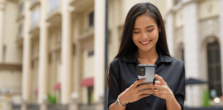 A woman with long dark hair smiles whilst looking at her mobile. She wears a black shirt and stands outdoors in front of a building with columns and arched windows. The background is softly blurred, highlighting her as the main focus.