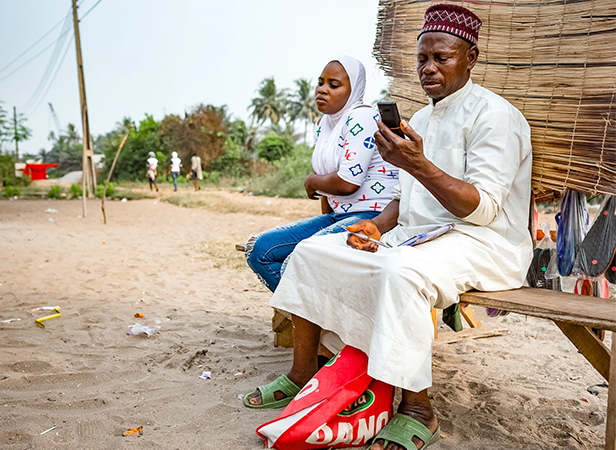 A man in traditional attire looks at his phone whilst sitting on a wooden bench outdoors; a woman in a white headscarf and colourful shirt sits beside him. Sandy ground, scattered litter, and people walking in the background with green trees.