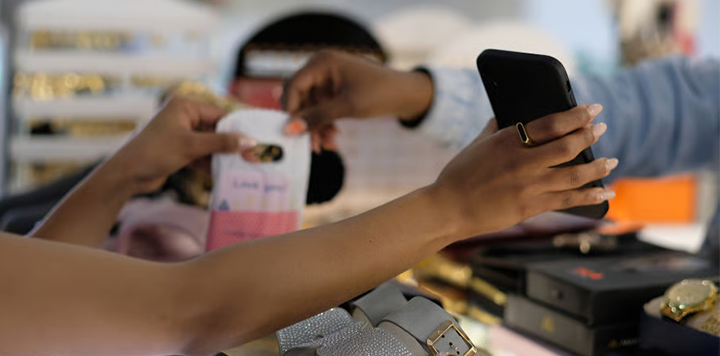 Two people’s hands interact over a retail counter filled with accessories. One person holds up a smartphone in a pink-trimmed case, whilst the other reaches towards it, both wearing rings and manicured nails.