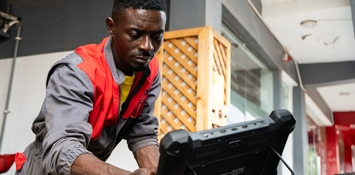 A man with short hair wears a grey and red boiler suit while focused on using a rugged tablet computer. He stands in a bright, modern workshop space with metal and wood structures in the background.